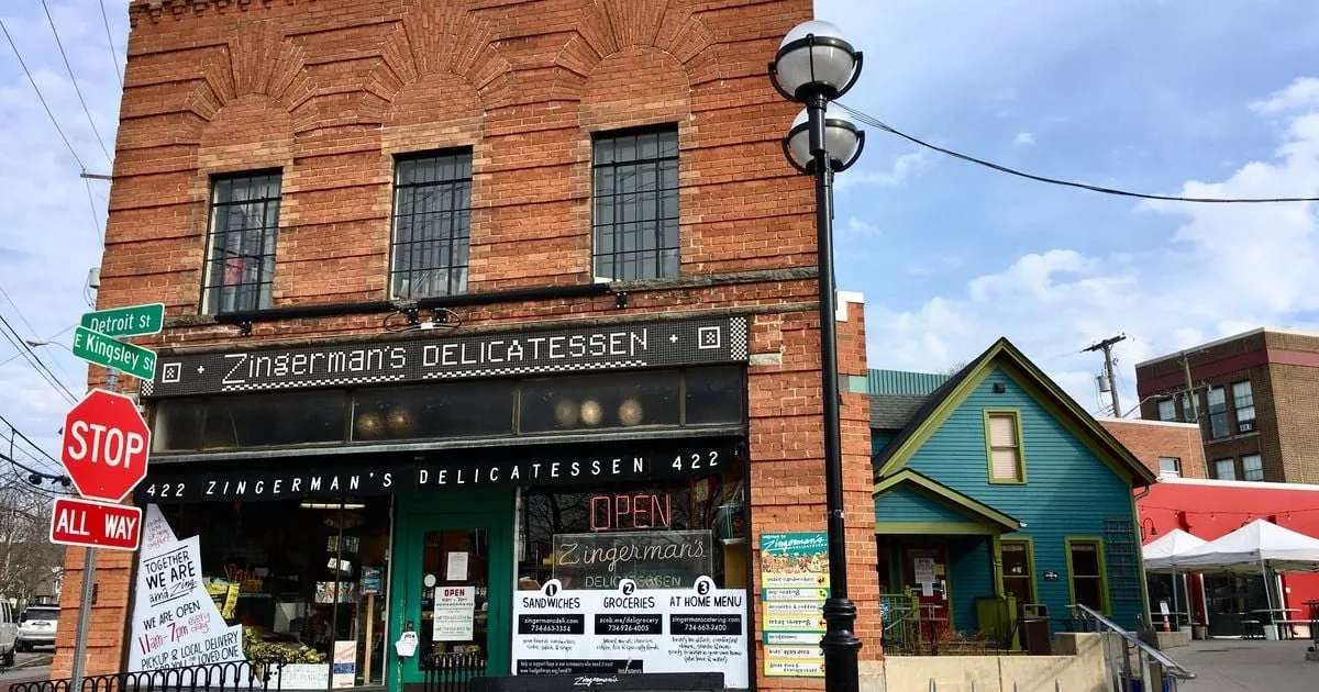 Exterior photo of Zingerman&rsquo;s Delicatessen in Ann Arbor, a brick corner storefront with a &lsquo;Zingerman&rsquo;s Delicatessen&rsquo; sign, street signs (Detroit St and E Kingsley St), a stop sign, and an &lsquo;OPEN&rsquo; window sign.